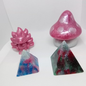 Peut inclure: Trois objets artisanaux en résine : un champignon rose à pois blancs, une créature rose ressemblant à un renard et une pyramide verte, bleue et rose.