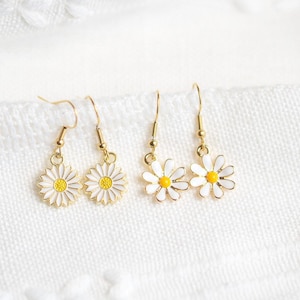 May include: Two pairs of daisy earrings with gold-colored hooks. The daisy pendants have white petals and yellow centers. The earrings are displayed on a white, textured surface.