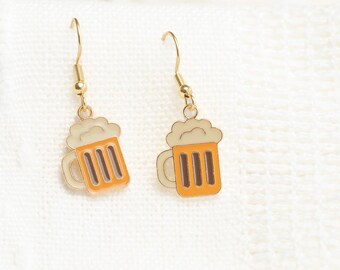 May include: A pair of gold-tone dangle earrings. Each earring features a miniature beer mug design with orange and brown details, topped with a white foam. The earrings are set against a white, textured background.