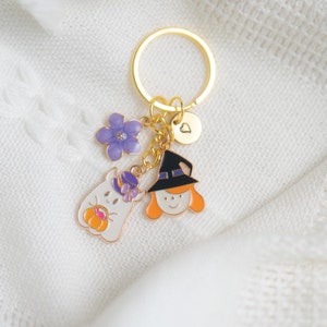 May include: A gold key chain with a purple flower charm, a witch charm, a ghost charm, and a heart charm. The charms are attached to a gold chain and a gold ring.