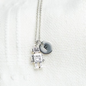 May include: A silver robot charm necklace with a silver letter 'F' charm. The necklace is on a white knitted surface.