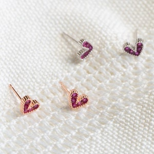 May include: Three pairs of small heart-shaped earrings with pink gemstones. The earrings are on a white fabric background.