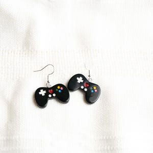 May include: A pair of black gaming controller earrings. Each earring features a controller shape with colorful buttons and a white cross button. The earrings hang from silver-colored hooks, set against a white background.