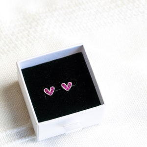 May include: A pair of heart-shaped earrings with pink rhinestones in a white box with a black interior.