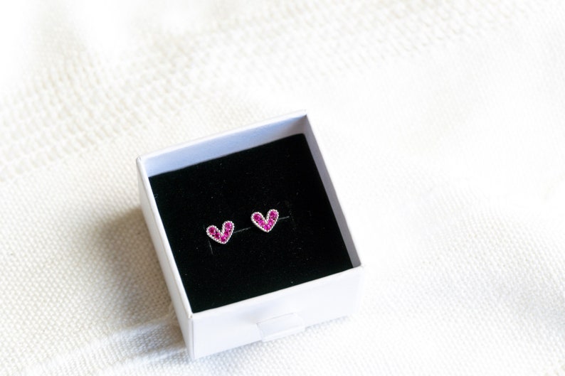 May include: A pair of heart-shaped earrings with pink rhinestones in a white box with a black interior.