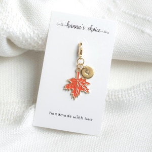 May include: A gold-toned metal charm featuring a bright orange maple leaf and a small circular disc with an engraved heart. The charm is attached to a white display card with the text hanna's choice and handmade with love printed on it.