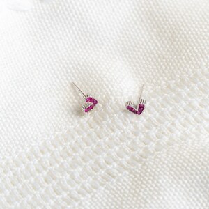 May include: A pair of silver heart-shaped earrings with pink gemstones. The earrings are on a white knitted surface.