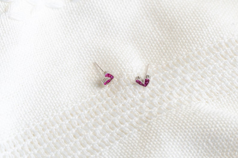 May include: A pair of silver heart-shaped earrings with pink gemstones. The earrings are on a white knitted surface.
