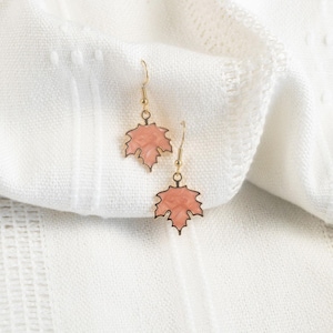 May include: A pair of leaf-shaped earrings with a gold-colored outline and ear hooks. The leaves are filled with a light peach color. The earrings are displayed on a white, textured fabric.