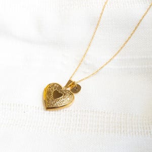May include: A gold heart-shaped locket necklace with a small gold initial charm. The necklace is on a white knitted surface.