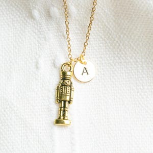 May include: Gold necklace with a nutcracker charm and a round pendant with the letter 'A' on it.