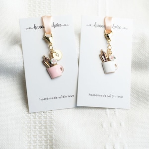 May include: Two gold keychains with a pink and white mug charm with pencils and flowers inside. The charms are attached to a gold chain with a heart charm. The keychains are on white cards with the text "hanna choice" and "handmade with love".