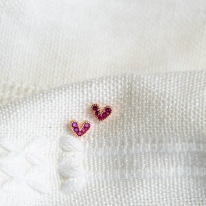 May include: A pair of small heart-shaped earrings with pink gemstones. The earrings are on a white fabric background.