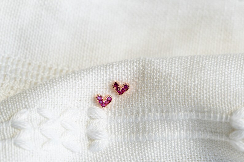 May include: A pair of small heart-shaped earrings with pink gemstones. The earrings are on a white fabric background.