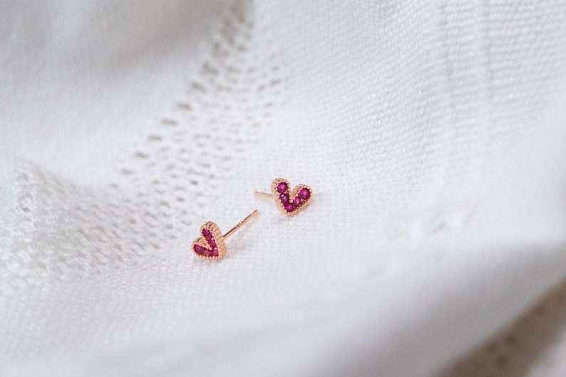 May include: A pair of rose gold heart-shaped earrings with small red gemstones.