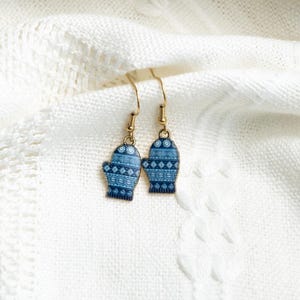 May include: A pair of blue mitten earrings with gold-colored hooks. The mittens have a patterned design in shades of blue and white. The earrings are displayed on a white, textured fabric.