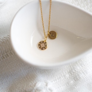May include: A gold necklace with a small heart charm and a larger charm with a star design and small clear stones. The necklace is displayed on a white ceramic dish.