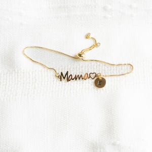 May include: Gold chain bracelet with a "Mama" charm and a heart. The bracelet also has a small charm with the letter "E" on it.