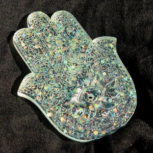 May include: A clear, iridescent resin hand-shaped incense burner with a detailed design. The burner is decorated with glitter and has a small hole in the center for burning incense.