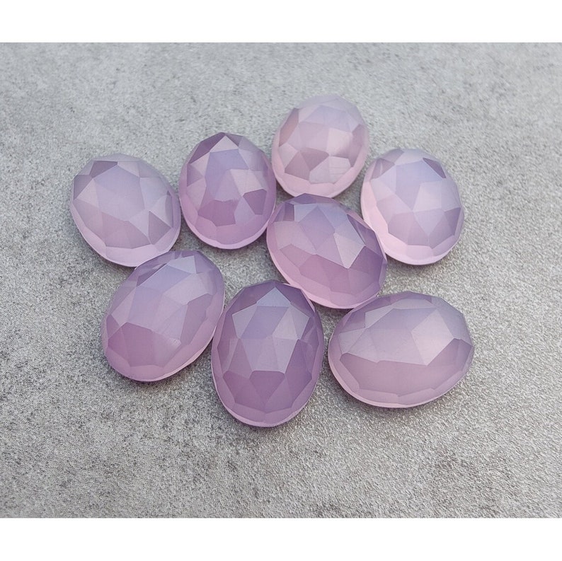 May include: Eight faceted oval-shaped gemstones in a pale lavender color. The stones are arranged in a cluster on a gray surface.