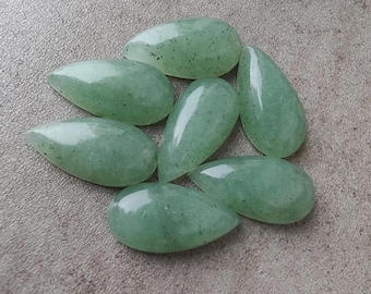 Op de afbeelding: Zeven gepolijste, druppelvormige groene aventurijn edelstenen. De stenen hebben een glad, glanzend oppervlak met kleine, donkere insluitsels. De edelstenen zijn gerangschikt op een neutraal, getextureerd oppervlak.