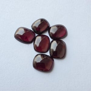 May include: A collection of six loose, cushion-cut garnet gemstones. Each stone features a deep, dark red color with a smooth, polished surface that reflects light, showcasing a rounded square shape with slightly curved edges and a glossy finish.