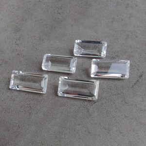 May include: Five clear, rectangular, faceted gemstones arranged in a scattered pattern. Each stone features a flat top surface with beveled edges, creating a transparent, glass-like appearance with sharp, defined corners and a polished finish.