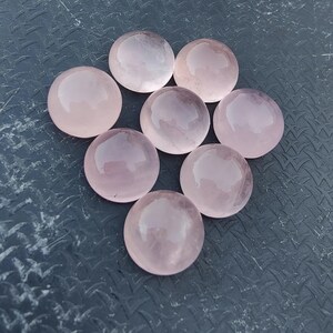 May include: Seven round, smooth, pink rose quartz stones. The stones are arranged in a circle on a textured grey surface.