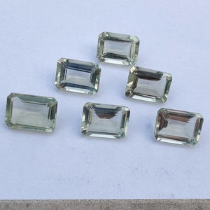 Pode incluir: Seis gemas de cor verde esmeralda cortadas em forma de esmeralda. As gemas são facetadas e têm uma aparência clara e transparente.