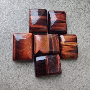 May include: Six polished, rectangular red tiger eye gemstones. The stones have a glossy finish and display a natural chatoyancy with bands of reddish-brown and gold hues. The gemstones are arranged on a gray surface.