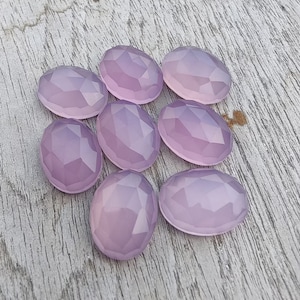 May include: Eight oval-shaped, faceted, light purple gemstones. The gemstones are arranged in a cluster on a light wood surface.