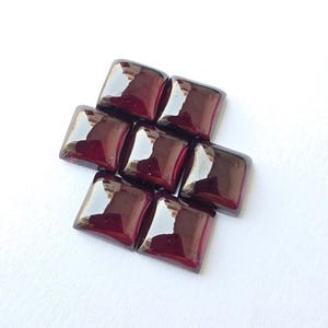 May include: A cluster of six square-cut, deep red garnet gemstones arranged in a compact formation. Each stone features a smooth, polished surface with reflective highlights, showcasing a rich, dark crimson hue and a uniform, flat-topped shape.