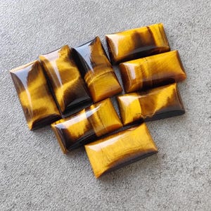 May include: Eight polished tiger's eye cabochons in various sizes and shapes, with a golden brown colour and black stripes. The stones are arranged in a heart shape on a grey surface.