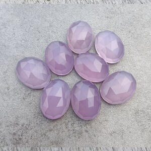 May include: Eight faceted oval-shaped gemstones in a pale lavender color. The stones are arranged in a loose circle on a gray surface.