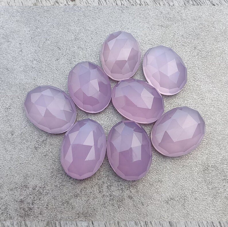 May include: Eight faceted oval-shaped gemstones in a pale lavender color. The stones are arranged in a loose circle on a gray surface.