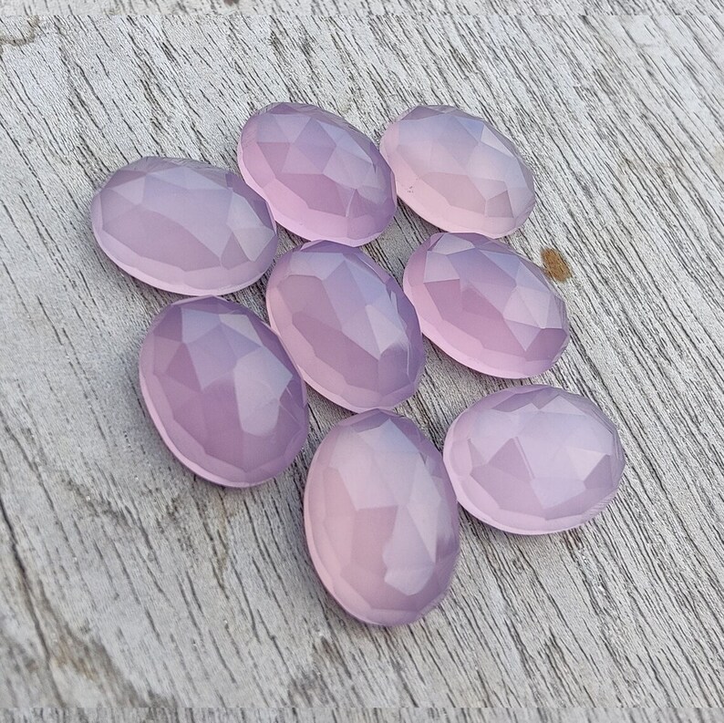 May include: Eight faceted oval-shaped gemstones in a pale lavender color. The stones are arranged in a loose cluster on a light wood surface.