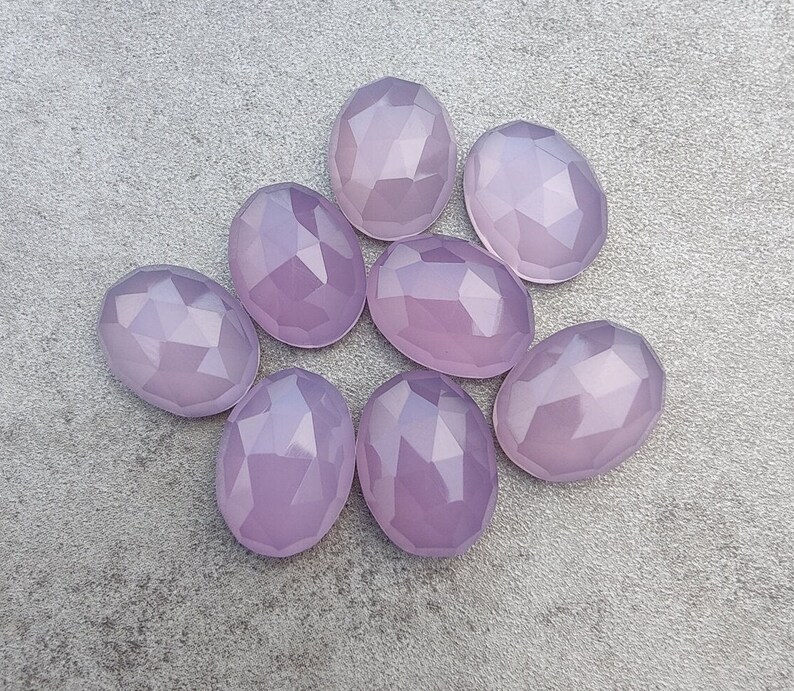 May include: Eight faceted oval-shaped gemstones in a pale lavender color. The stones are arranged in a loose circle on a gray background.