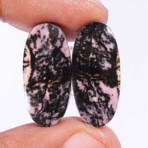 May include: Two oval gemstones with a polished surface. The stones display a mix of black and pink hues, with a marbled pattern. The gemstones are held between fingers, showing their size and shape. The stones are likely used for jewellery making.