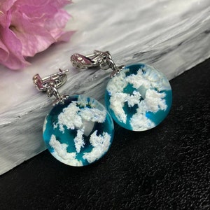 Puede incluir: Un par de pendientes colgantes de plata con resina azul e inclusiones blancas en forma de nube. Los pendientes son redondos y tienen un cierre de gancho de plata.