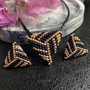 May include: A black and gold beaded triangle necklace and earrings set. The necklace has a black leather cord and the triangle pendant is made of black and gold beads. The earrings are also triangle shaped and made of black and gold beads.