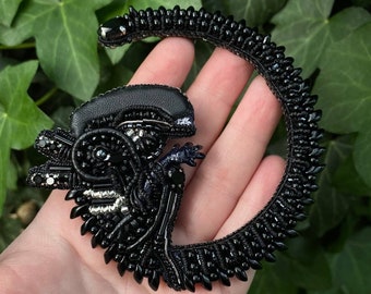 May include: A black beaded brooch shaped like a xenomorph creature with a long, curved tail. The piece is crafted with intricate black beads, sequins, and metallic accents, featuring a smooth leather-like head section and a textured, spiky body.