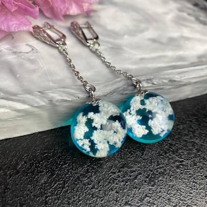 Puede incluir: Un par de pendientes de aro de plata con un colgante de esfera de resina azul y blanca. La esfera contiene motas blancas que se asemejan a nubes.