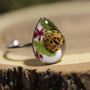 Könnte beinhalten: Ein silberfarbener Ring mit einer tropfenförmigen Fassung, die einen gelben Marienkäfer mit schwarzen Punkten, grüne Blätter und kleine violette Blüten in einem klaren, glatten, harzähnlichen Material enthält.