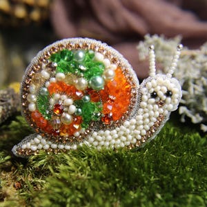 May include: A handcrafted snail brooch, meticulously beaded with white, orange, green, and pearl-colored beads. The snail's shell features a spiral design, and the body is covered in small white beads. The brooch is set against a backdrop of green moss.