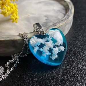 Puede incluir: Un collar de cadena plateada con un colgante en forma de corazón. El colgante está lleno de resina azul transparente y flores secas blancas.