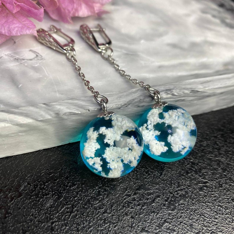 Puede incluir: Un par de pendientes de aro de plata con un colgante de esfera de resina azul y blanca. La esfera contiene motas blancas que se asemejan a nubes.