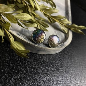 Peut inclure: Une paire de boucles d'oreilles rondes &agrave; clous iris&eacute;es avec un motif textur&eacute; en forme d'&eacute;cailles. Les boucles d'oreilles sont sur une surface en marbre blanc.