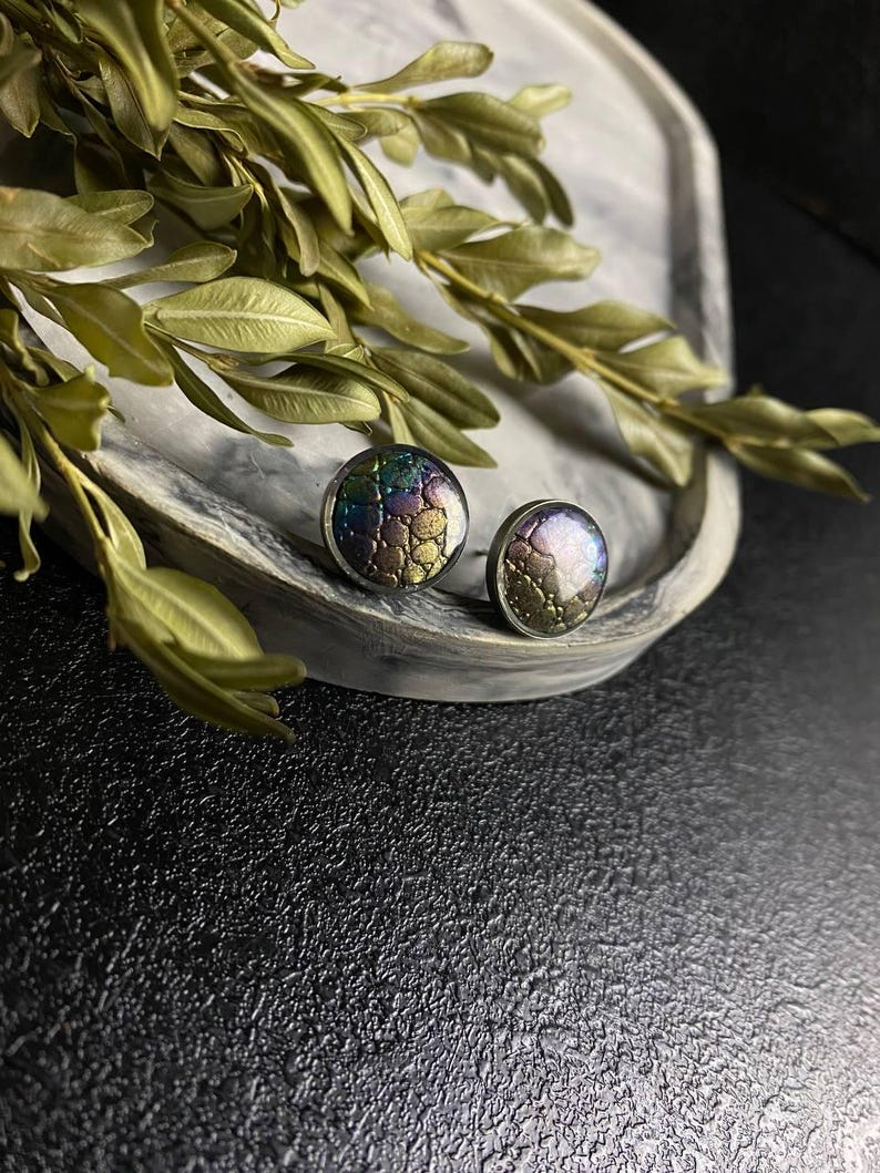 Peut inclure: Une paire de boucles d'oreilles rondes &agrave; clous iris&eacute;es avec un motif textur&eacute; en forme d'&eacute;cailles. Les boucles d'oreilles sont sur une surface en marbre blanc.