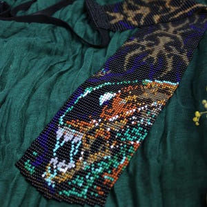 Puede incluir: Una diadema de cuentas con un diseño detallado en negro, azul, naranja y verde. La diadema está sobre una tela verde oscuro, con una cinta negra adjunta. El diseño parece ser un animal o criatura estilizada.