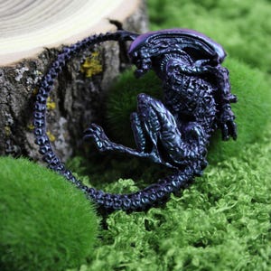 May include: A detailed, iridescent purple and blue Xenomorph figurine, posed on a bed of vibrant green moss. The alien creature is curled up, showcasing its textured exoskeleton and elongated tail. A slice of a tree trunk is in the background.